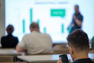 Blurred view of a lecture with a man focusing on his smartphone in the foreground.