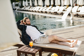Businessman working remotely by the poolside with laptop and cocktail