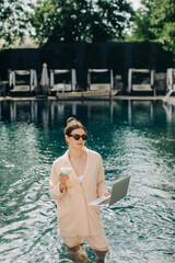 Businesswoman working on laptop and holding cocktail in pool