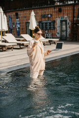 Businesswoman working remotely from swimming pool with laptop and cocktail