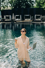 Businesswoman working remotely from swimming pool with laptop and cocktail