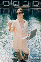 Businesswoman working remotely from the pool with laptop and cocktail