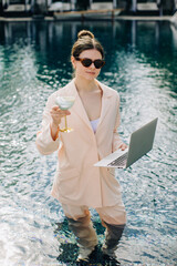 Businesswoman working remotely from swimming pool with laptop and cocktail