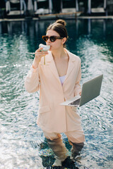 Businesswoman working remotely from swimming pool drinking cocktail