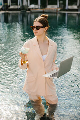 Businesswoman working remotely from swimming pool with laptop and cocktail