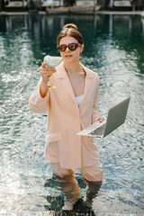 Businesswoman working remotely from swimming pool with laptop and cocktail