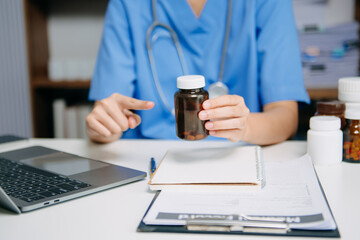 Healthcare professional pouring medication into hand while working with laptop and patient files,...