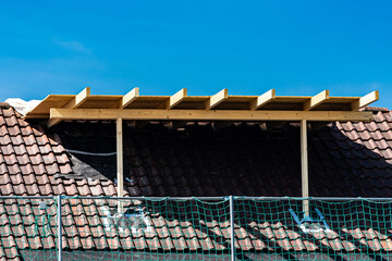 Fototapeta premium A wooden framework is being constructed on a slanted roof at a residential site. The blue sky serves as a backdrop while scaffolding and construction materials are visible nearby.