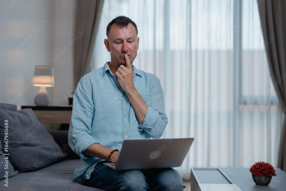 Poster thoughtful businessman is sitting on the sofa with a laptop on his lap and touching his chin with a 