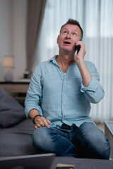 Mature man sitting on a sofa at home, making a phone call and displaying a worried expression while...