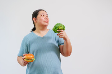 A plus-size Asian overweight woman holding both burger and broccoli, choice between healthy and...