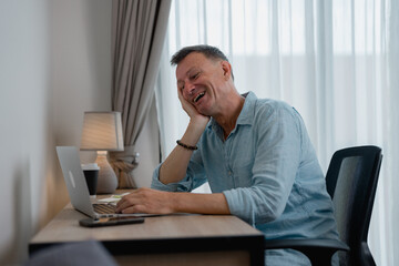 Fototapeta premium Mature businessman is sitting at his desk, working on his laptop and laughing, enjoying the benefits of working remotely in a comfortable and relaxed setting