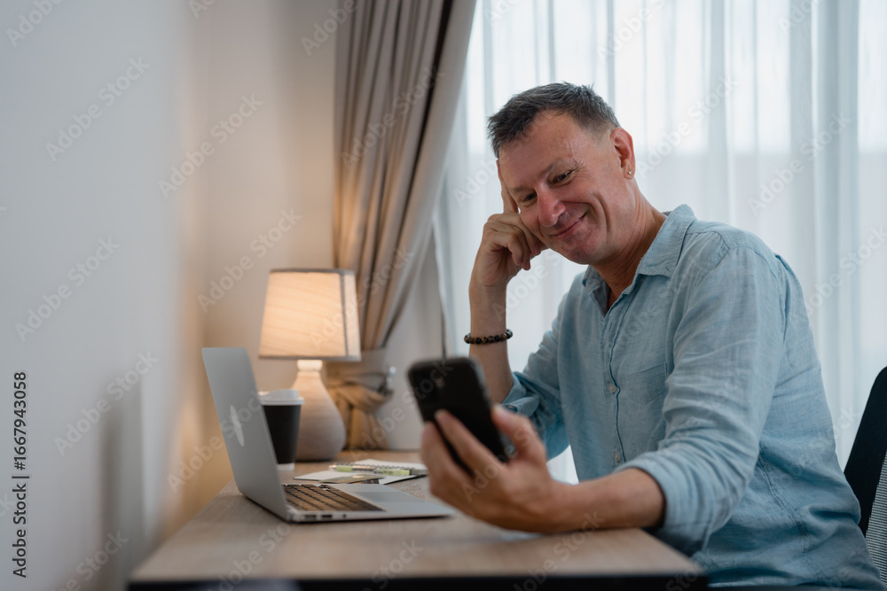 Poster mature businessman sitting at a desk in a home office, smiling while using a smartphone. laptop and 