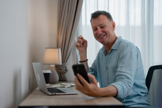 Smiling mature businessman having video call on smartphone and gesturing while sitting at desk with laptop and takeaway coffee in home office