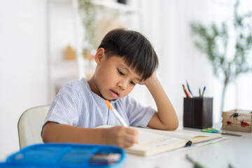 Asian boy studying with pencil at home smiling writing with notebook and school supplies, online...