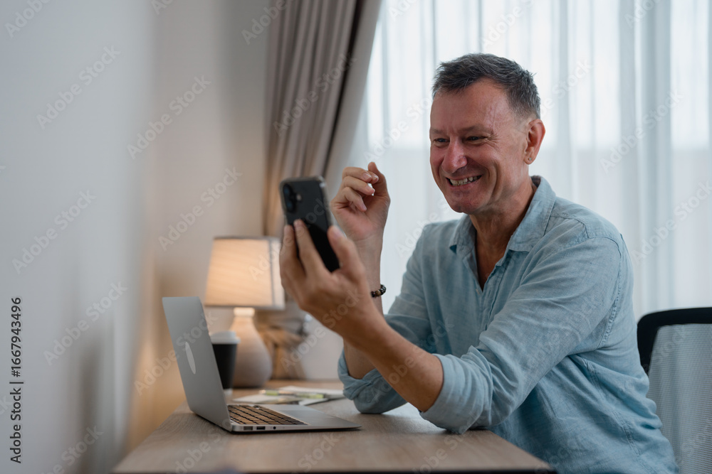 Poster smiling mature man engaging in a video call while seated at a desk with a laptop, enjoying the exper