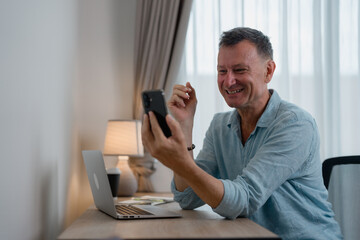 Smiling mature man engaging in a video call while seated at a desk with a laptop, enjoying the experience of online communication in a comfortable home office setting
