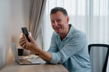 Mature businessman sitting at a desk in a home office, displaying a smartphone with an excited...