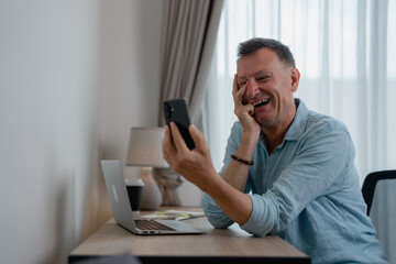 Grey-haired businessman sitting at his desk in a light blue shirt, laughing joyfully during a video...