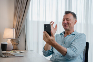 Smiling mature man waving hand while having video conference using mobile phone at desk in bright modern apartment, enjoying online communication