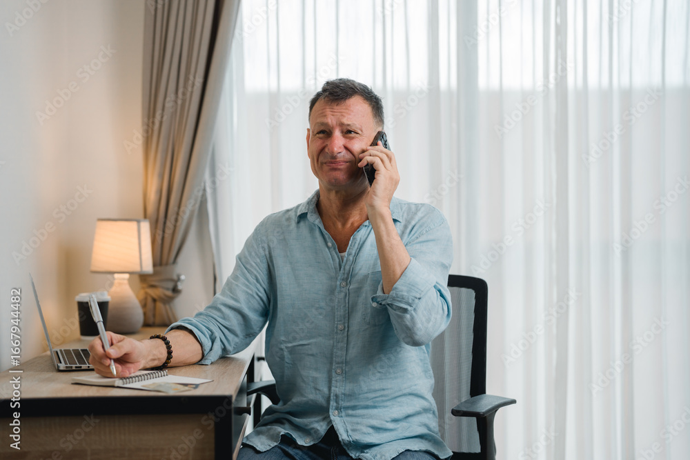 Poster mature businessman talking on mobile phone and taking notes in notebook while sitting at desk and wo
