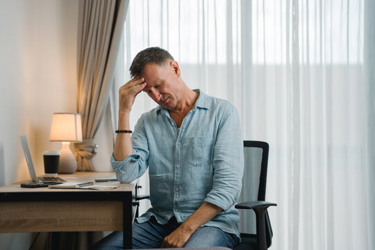 Businessman suffering from headache, migraine or stress sitting at work desk with laptop, he is massaging his head while working from home in his home office