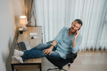 Relaxed businessman is talking on phone with feet up on desk enjoying remote work in home office, he's holding a pen in his hand and smiling