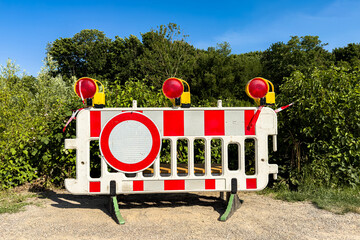 A traffic barrier with red and white stripes stands blocking a dirt road. Bright warning lights top...