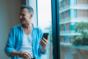Happy middle-aged man holding mobile phone standing next to a window in his modern apartment,...