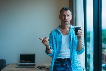Mature man with blue shirt and jeans drinking coffee from disposable cup and pointing with finger to the side while looking away near a window in his home office