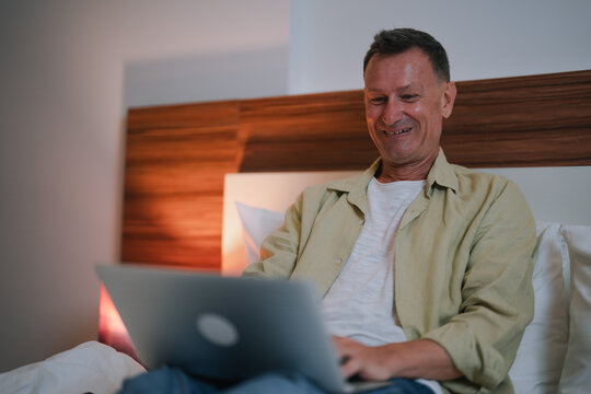 Happy mature man working or using social media on his laptop while sitting comfortably in bed at night, enjoying the convenience of technology in his bedroom