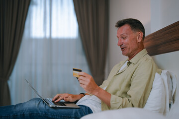 Happy mature man lounging on a bed in a hotel room, using a laptop and credit card for online...