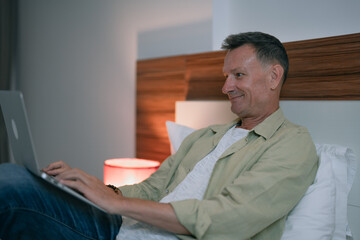 Mature man lying in bed at night, smiling while using his laptop, enjoying the comfort of his hotel room or home, and engaging in remote work with ease and productivity