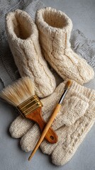 Knitted cream-colored slippers, gloves, and paintbrushes on gray surface