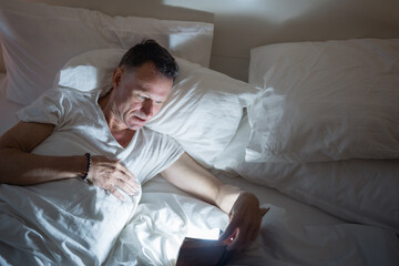 Senior man lying in bed at night, reading a book with a flashlight under the covers, struggling to...