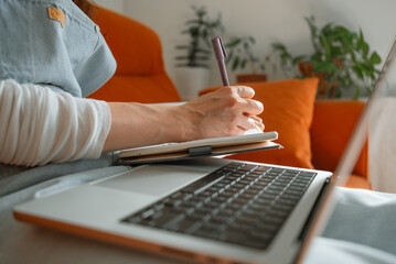 Woman sitting on a comfortable sofa, using laptop and writing notes in notebook, working from home,...