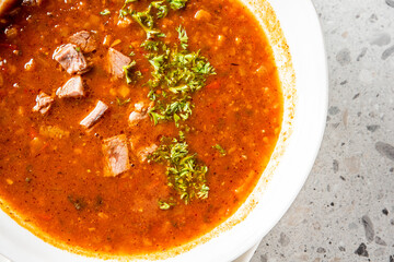 Close-up of hearty tomato-based soup with beef chunks and fresh parsley garnish, served in a white bowl on a speckled countertop. Rich, rustic, and homemade appearance.