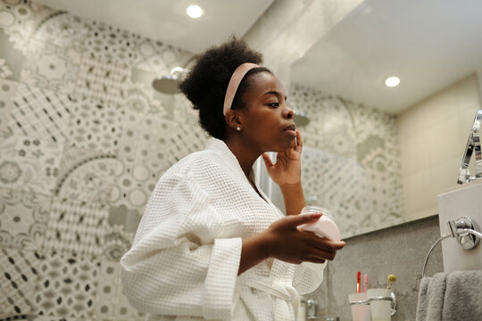 Low angle shot of beautiful young African American woman wearing white bath robe standing in front of mirror in modern bathroom applying nourishing cream on face