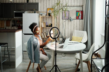 Horizontal long shot of stylish young woman with eye patches on face wearing casual clothes sitting at table in loft room at home taking selfie shots