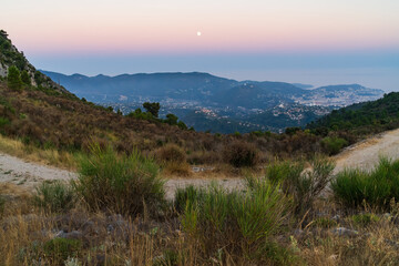 Serene mountain landscape with a hiking trail overlooking the coastal city of Nice under a rising full moon at twilight.