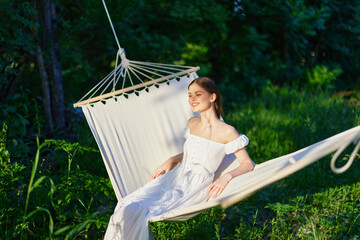 Young woman relaxing in a hammock in a lush green environment, wearing a white sundress, expressing...