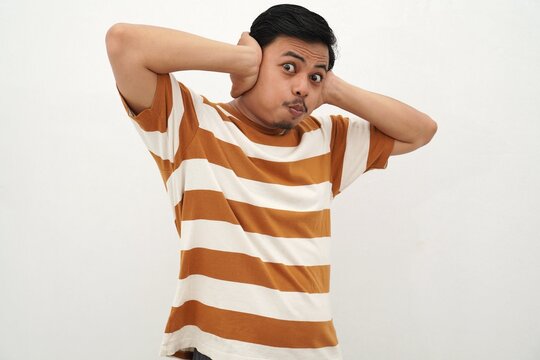 Young Asian man in white and brown striped t-shirt covering his ears with an annoyed expression showing frustration or stress against a white background