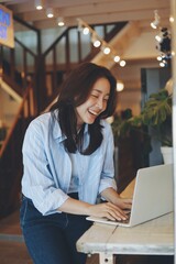Cheerful Asian woman smiling and laughing while using laptop at modern cafe, representing freelance lifestyle, digital nomad, and online business success.