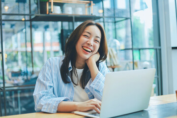 Cheerful Asian woman smiling and laughing while using laptop at modern cafe, representing freelance...