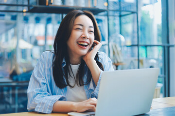 Cheerful Asian woman smiling and laughing while using laptop at modern cafe, representing freelance...