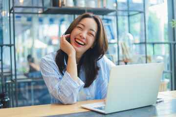 Cheerful Asian woman smiling and laughing while using laptop at modern cafe, representing freelance...