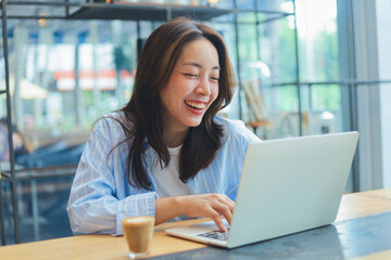 Cheerful Asian woman smiling and laughing while using laptop at modern cafe, representing freelance...
