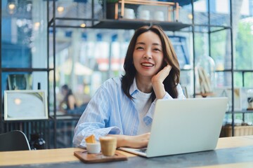 Cheerful Asian woman smiling and laughing while using laptop at modern cafe, representing freelance...