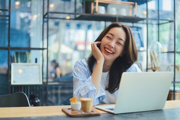 Cheerful Asian woman smiling and laughing while using laptop at modern cafe, representing freelance...