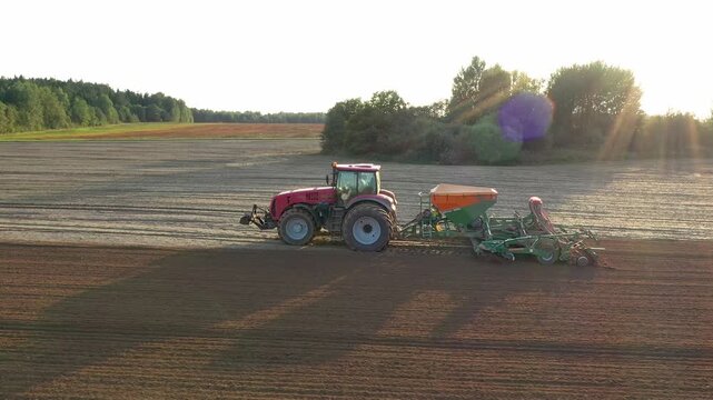 Modern tractor sowing cereal seeds rye wheat barley maize across fertile farmland. Rural machinery working on field. Aerial view. Farmer plowing agricultural field at sunset shines background